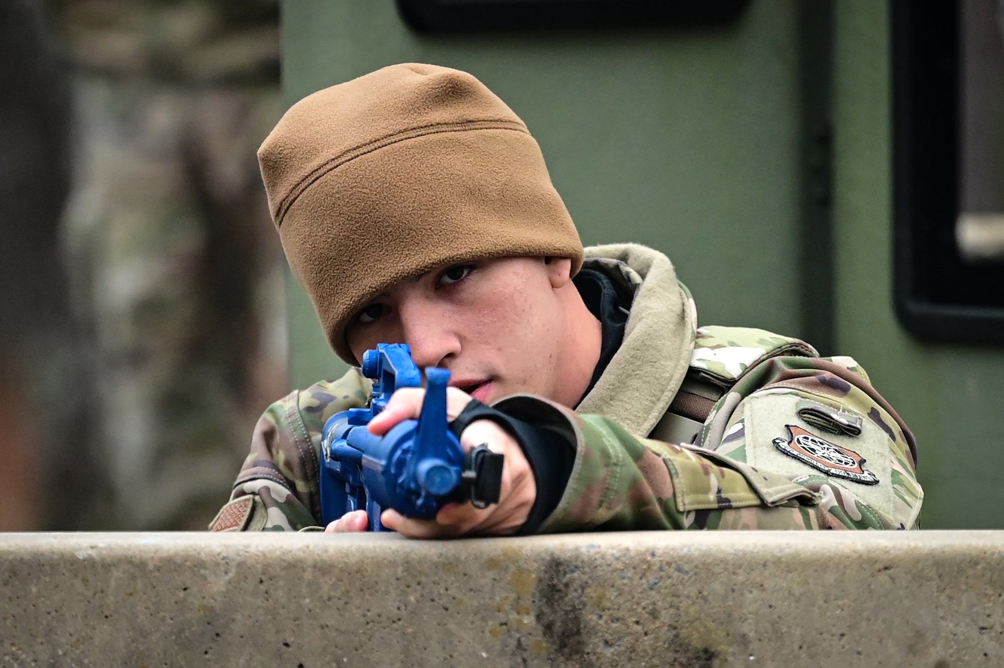 An Airman stands over a barricade with a blue training assault rifle.