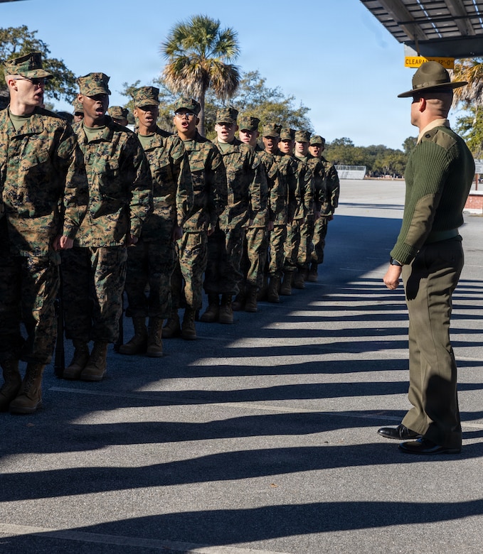 Ten people in camouflage military uniforms stand in line outdoors as a man in a formal military uniform observes them.