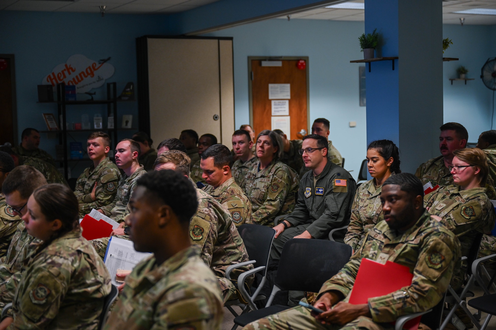 Airmen sit in a briefing room during an exercise. Some members have red folders in their hands.