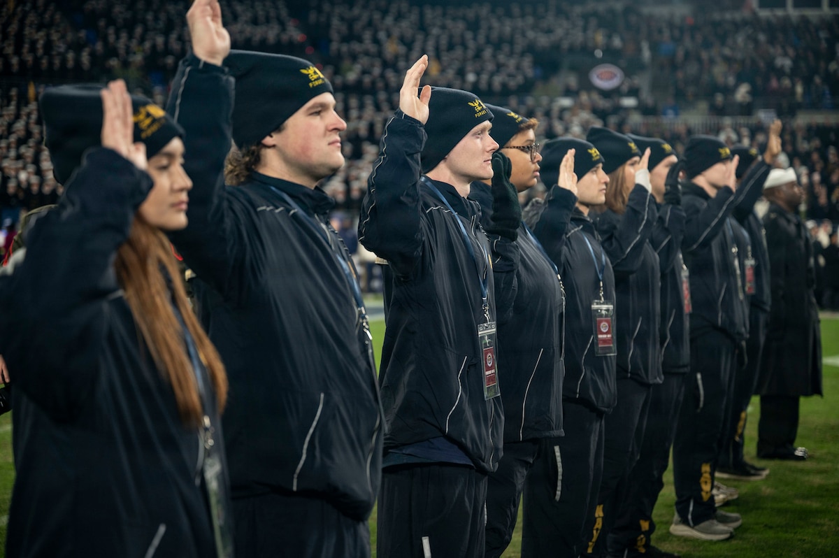 About a dozen people in winter athletic clothing stand on a field of grass, raising their right hands.