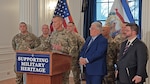 white men in military uniform stand at and around a podium next to a civilian man in a blue suit. indoors.