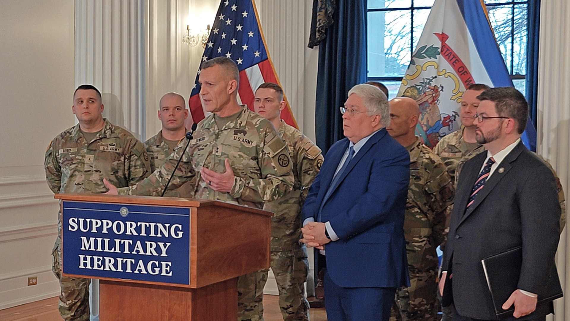 white men in military uniform stand at and around a podium next to a civilian man in a blue suit. indoors.