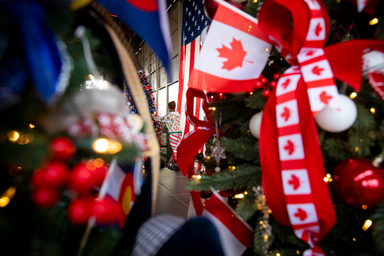 A woman in Christmas attire hangs decorations on a tree. There are decorations out of focus in the foreground.