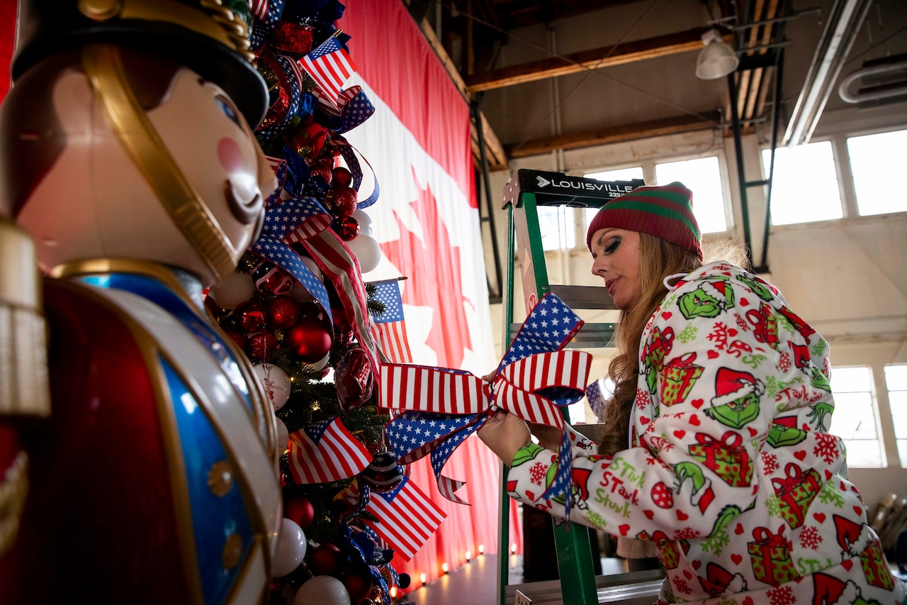A woman in Christmas attire hangs decorations while standing on a ladder.
