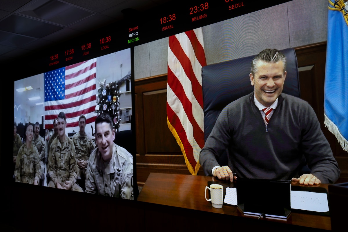 Two large video screens mounted on the wall of a large conference room. The left screen shows a group of service members in camouflage military uniforms sitting around a conference table. The right screen shows a man in business attire sitting at the head of a long conference table; behind him are an American flag and a blue flag with an eagle in the center.