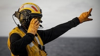 A sailor in yellow flight deck gear signals to aircraft on the deck of a large aircraft carrier.