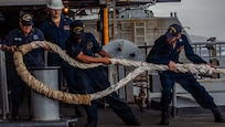 A group of sailors pulls a thick mooring line on the fantail of a large aircraft carrier.