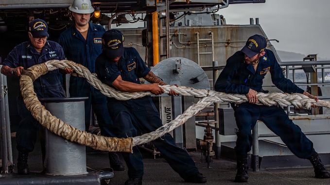 A group of sailors pulls a thick mooring line on the fantail of a large aircraft carrier.