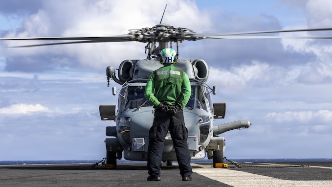 A sailor stands on the flight deck of an aircraft carrier next to an MH-60R Sea Hawk helicopter.