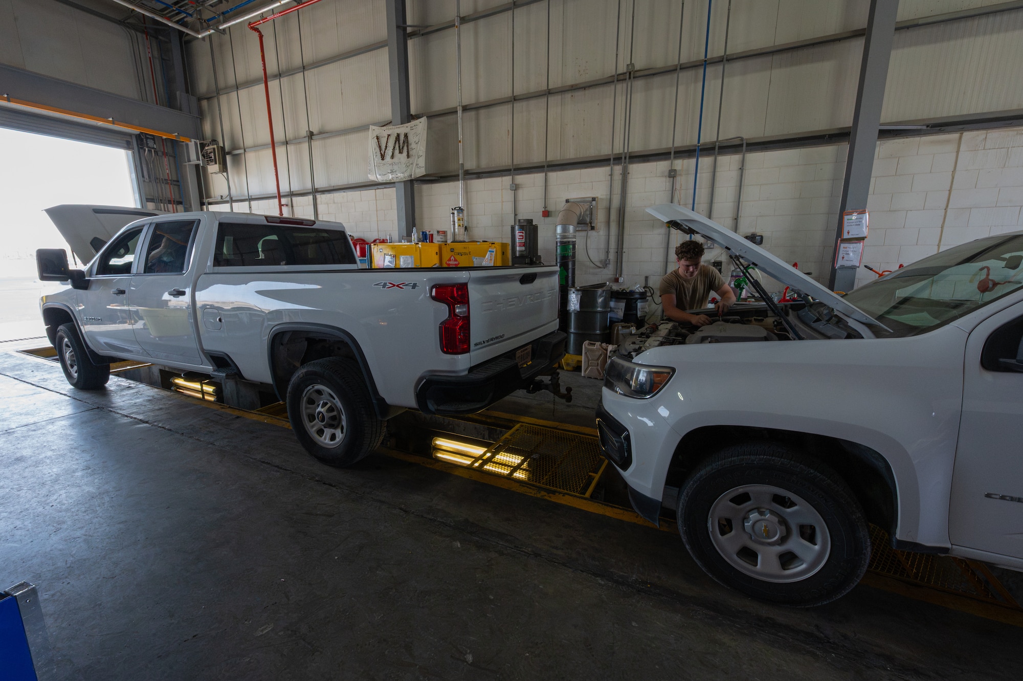Two trucks in a garage, one's hood is open and a vehicle maintenance person is working on it