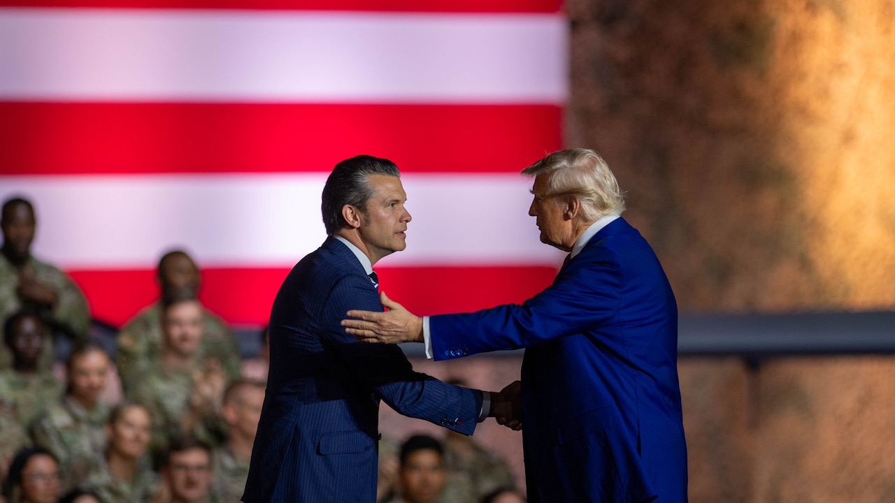 President Donald J. Trump shakes hands with Defense Secretary Pete Hegseth in front of a seated audience of troops and a U.S. flag.