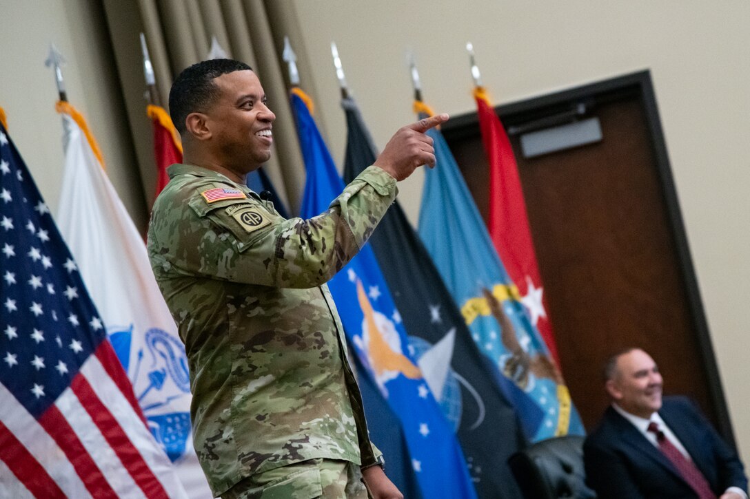 A man wearing a camouflage military uniform stands indoors on a stage smiling and pointing toward the audience.