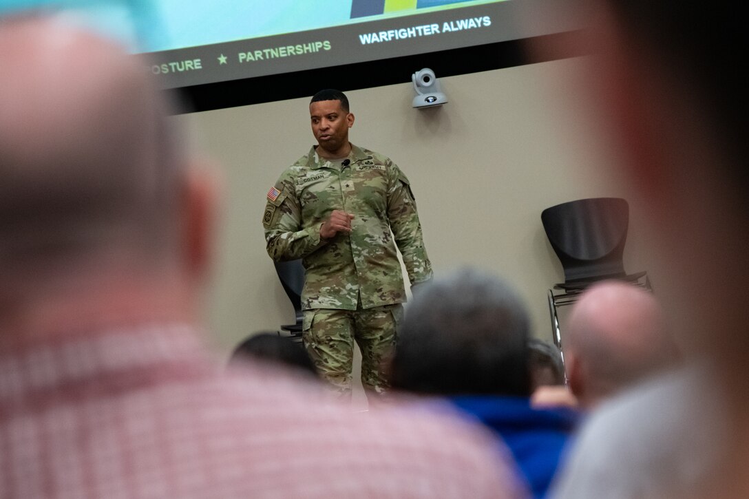 A man wearing a camouflage military uniform stands indoors on a stage with a large display screen behind him speaking to a large audience.