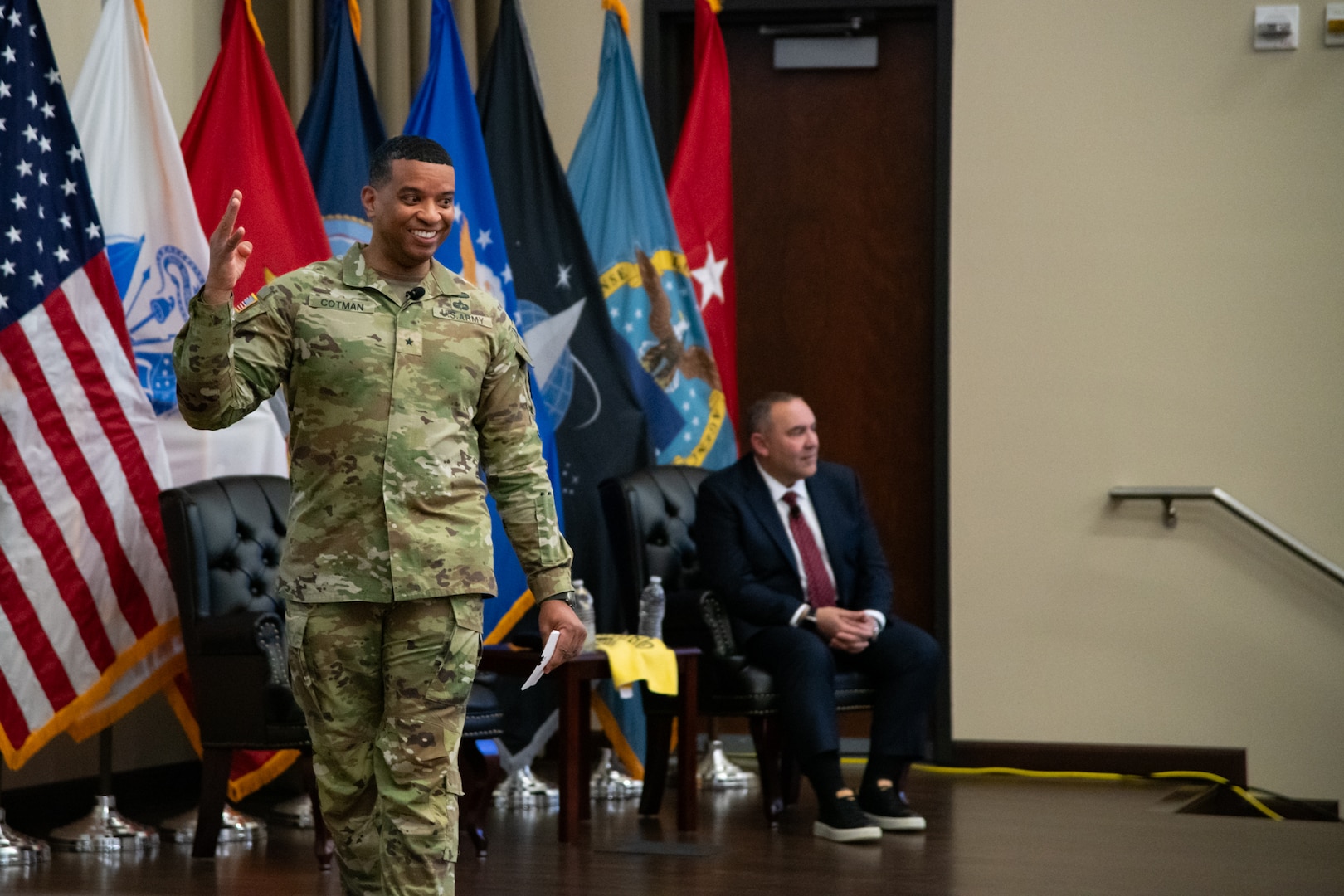 A man wearing a camouflage military uniform walks across a stage indoors while smiling and gesturing toward the audience.