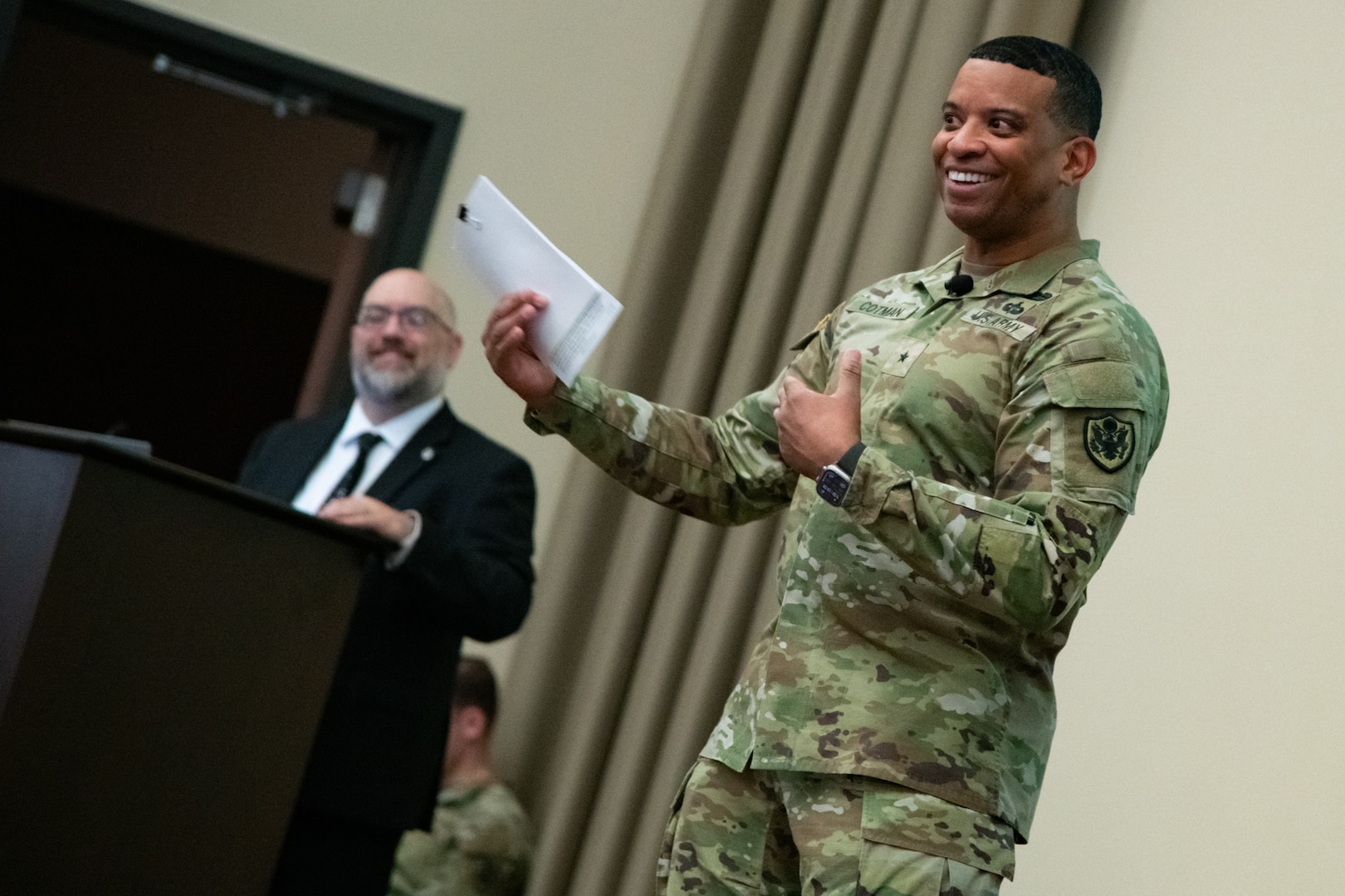 A man wearing a camouflage military uniform stands indoors on a stage gesturing to the audience and smiling.