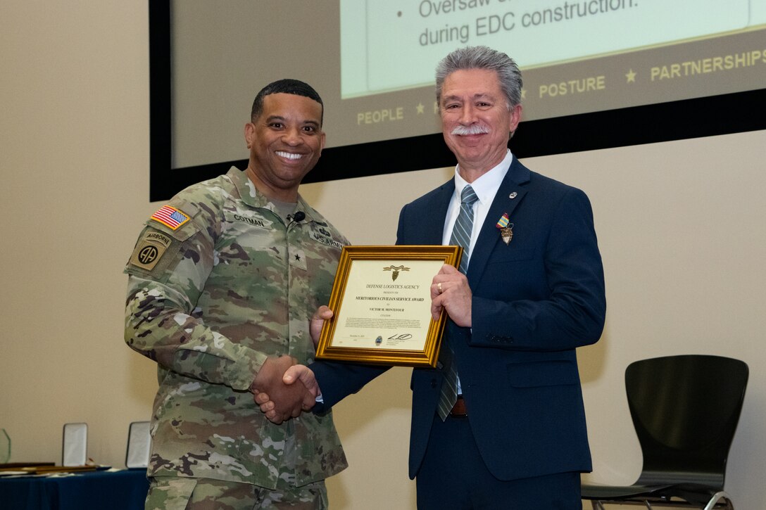 A man wearing a camouflage military uniform shakes hands with a man wearing a suit and holding a framed certificate. They are smiling and looking into the camera.