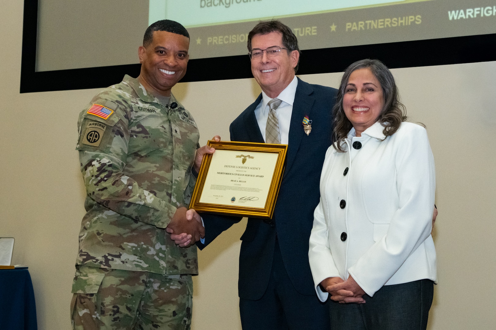 A man wearing a camouflage military uniform shakes hands with a man wearing a suit and holding a framed certificate. There is a woman standing next to the man in the suit. They are all smiling and looking into the camera.