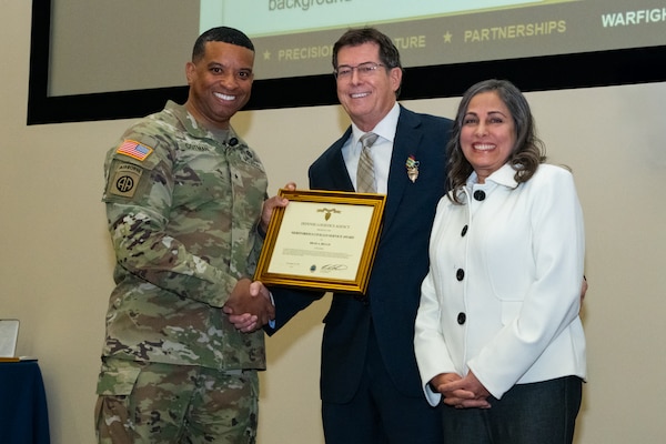 A man wearing a camouflage military uniform shakes hands with a man wearing a suit and holding a framed certificate. There is a woman standing next to the man in the suit. They are all smiling and looking into the camera.