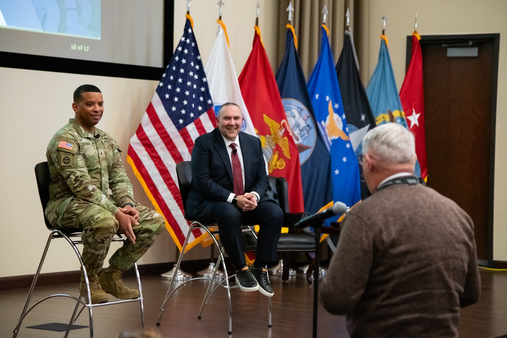 Two men, one wearing a suit and one wearing a camouflage military uniform, sit indoors on stools on a stage looking toward a man in the audience who is facing them and speaking into a microphone on a microphone stand.