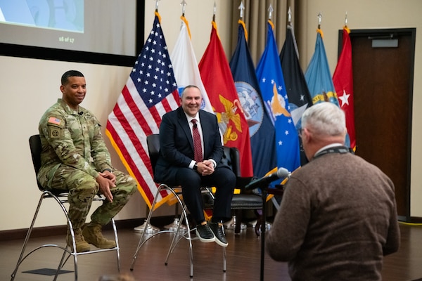 Two men, one wearing a suit and one wearing a camouflage military uniform, sit indoors on stools on a stage looking toward a man in the audience who is facing them and speaking into a microphone on a microphone stand.