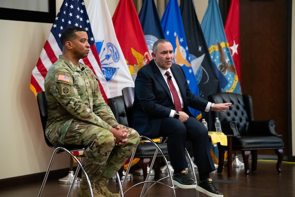Two men sit on stools on a stage, one wearing a suit and one wearing a camouflage military uniform.