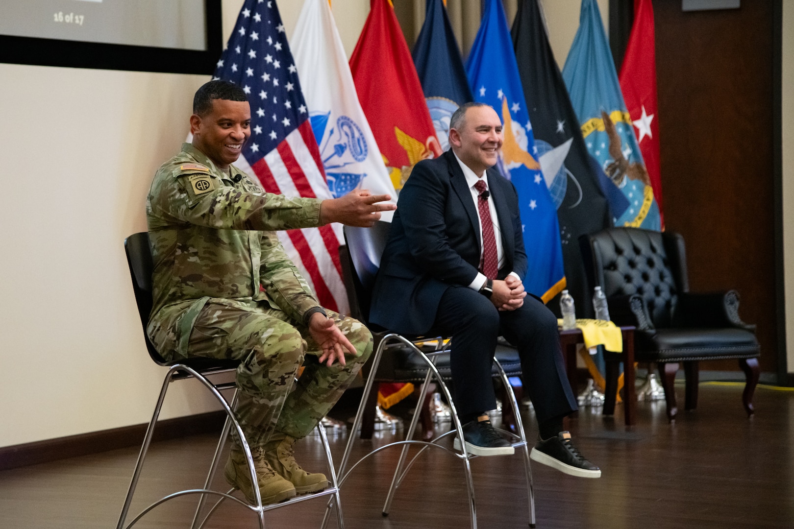 Two men sit on stools on a stage, one wearing a suit and one wearing a camouflage military uniform. The man in the uniform is gesturing toward the audience and smiling.