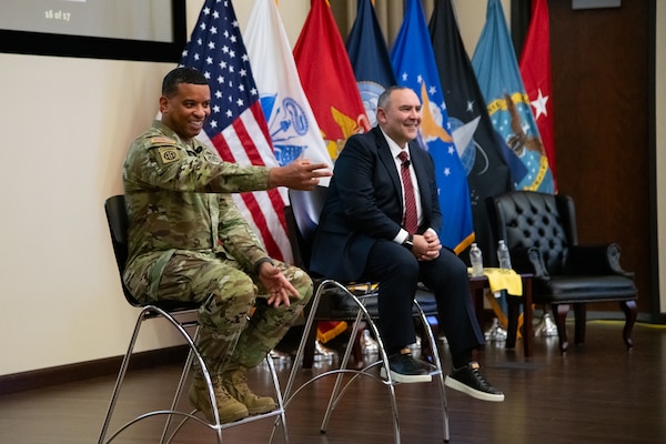 Two men sit on stools on a stage, one wearing a suit and one wearing a camouflage military uniform. The man in the uniform is gesturing toward the audience and smiling.