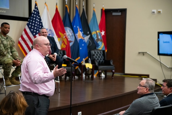A man stands in front of an indoor stage speaking into a microphone on a microphone stand.