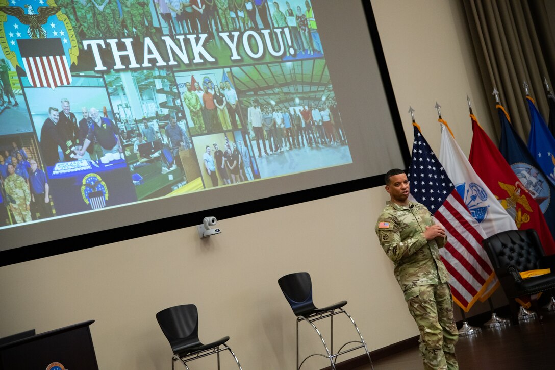 A man wearing a camouflage military uniform stands indoors on a stage with a screen behind him that reads "thank you."