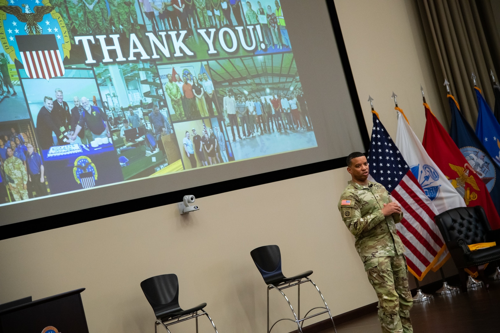 A man wearing a camouflage military uniform stands indoors on a stage with a screen behind him that reads "thank you."