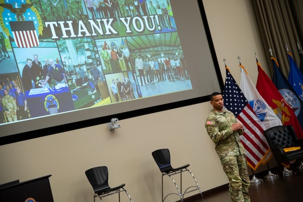 A man wearing a camouflage military uniform stands indoors on a stage with a screen behind him that reads "thank you."
