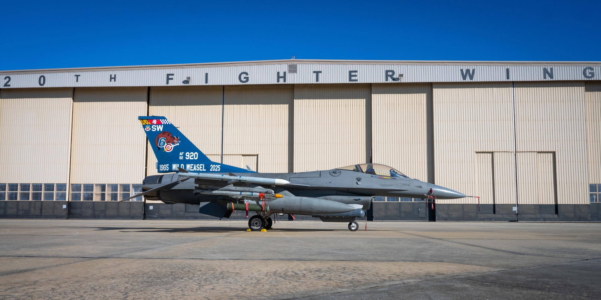 A fighter jet with a blue tail is parked in front of a hangar that says 20th Fighter Wing at the top