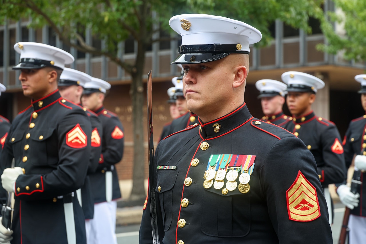 A Marine wearing black formal attire and a white hat stands at attention among other Marines outdoors.
