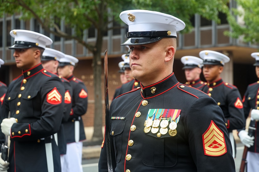 A Marine wearing black formal attire and a white hat stands at attention among other Marines outdoors.