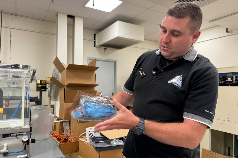A man holds a plastic pouch in a room filled with machines and cardboard boxes.