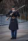 An Army Soldier wearing ceremonial service dress with sunglasses is standing at attention holding a ceremonial rifle diagonally in front of his chest.