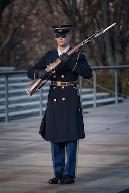 An Army Soldier wearing ceremonial service dress with sunglasses is standing at attention holding a ceremonial rifle diagonally in front of his chest.