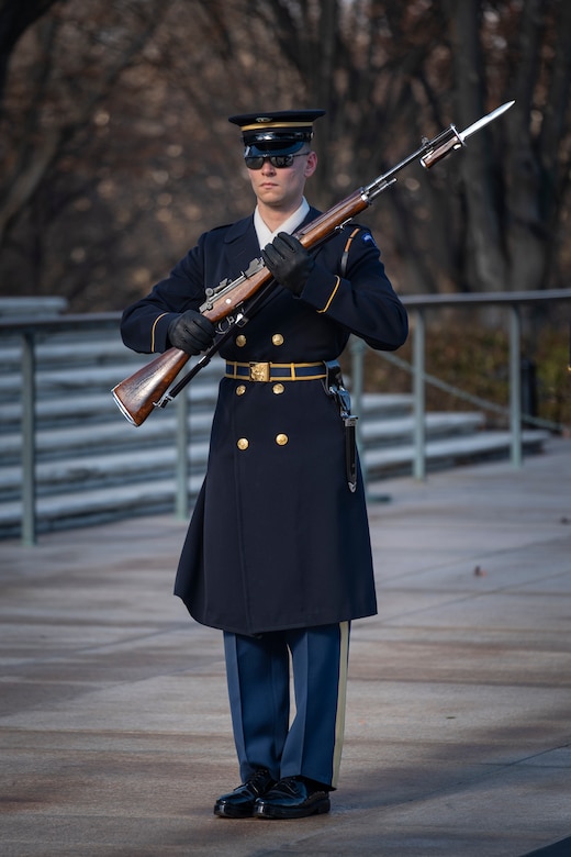 An Army Soldier wearing ceremonial service dress with sunglasses is standing at attention holding a ceremonial rifle diagonally in front of his chest.