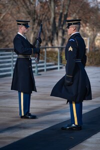Two Army Soldiers wearing ceremonial uniforms with overcoats are standing at attention, one standing on a black mat while the other is holding a ceremonial rifle in front of his body.