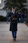 An Army Soldier wearing ceremonial service dress with sunglasses is marching toward the camera while holding a ceremonial rifle diagonally in front of his chest.
