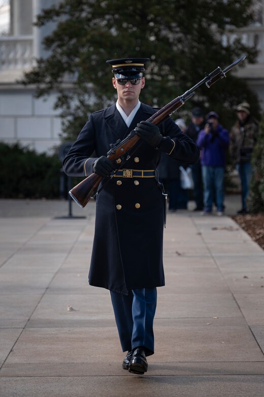 An Army Soldier wearing ceremonial service dress with sunglasses is marching toward the camera while holding a ceremonial rifle diagonally in front of his chest.
