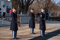 The Army Soldiers are standing in a triangle formation, two holding ceremonial rifles in front of their bodies, and one standing on a long black mat at the Tomb of the Unknown Soldier in Arlington National Cemetery.
