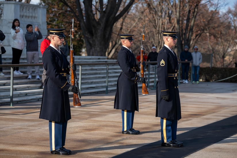 The Army Soldiers are standing in a triangle formation, two holding ceremonial rifles in front of their bodies, and one standing on a long black mat at the Tomb of the Unknown Soldier in Arlington National Cemetery.