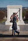 An Army Soldier wearing ceremonial service dress with sunglasses is standing at attention holding a ceremonial rifle diagonally in front of his chest.