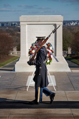 An Army Soldier wearing ceremonial service dress with sunglasses is standing at attention holding a ceremonial rifle diagonally in front of his chest.