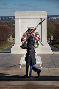 An Army Soldier wearing ceremonial service dress with sunglasses is standing at attention holding a ceremonial rifle diagonally in front of his chest.