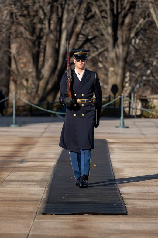An Army Soldier wearing ceremonial service dress with sunglasses is marching down a black mat at the Tomb of the Unknown Soldier in Arlington National Cemetery toward the camer while carrying a ceremonial rifle on his right shoulder.
