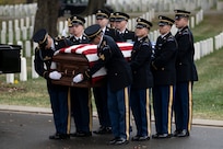 Eight Army soldiers wearing ceremonial dress uniforms as carrying a U.S. flag-draped casket during a funeral at a cemetery.