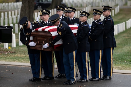 Eight Army soldiers wearing ceremonial dress uniforms as carrying a U.S. flag-draped casket during a funeral at a cemetery.