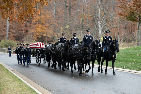A U.S. Army caisson is moving down a street during a funeral with four Army Soldiers riding horses and other soldiers marching alongside.